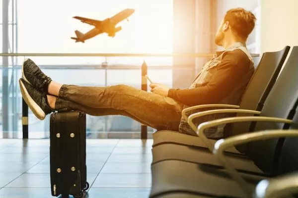 Jemand sitzt am Flughafen im Wartebereich, hat die Füße auf den Koffer hochgelegt und schaut im Sonnenuntergang einem abhebenden Flugzeug zu