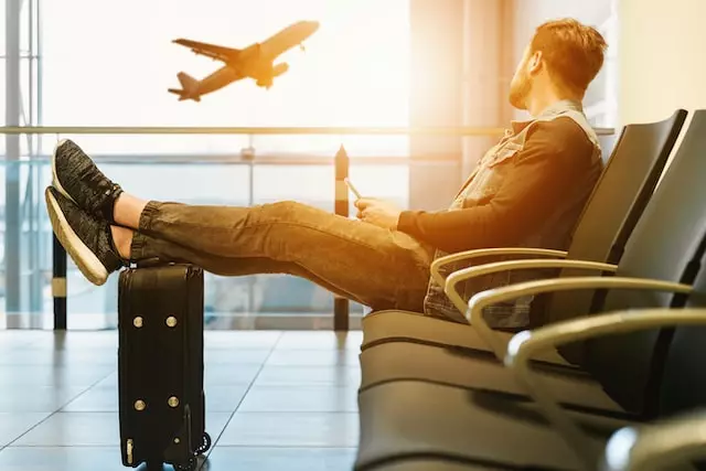 Jemand sitzt am Flughafen im Wartebereich, hat die Füße auf den Koffer hochgelegt und schaut im Sonnenuntergang einem abhebenden Flugzeug zu