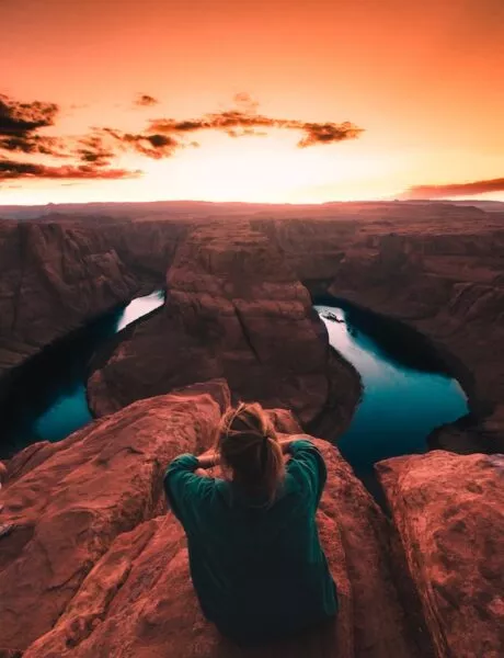 Eine Frau sitzt mit dem Rücken zur Kamera oben auf einem Canyon und schaut in den Sonnenuntergang