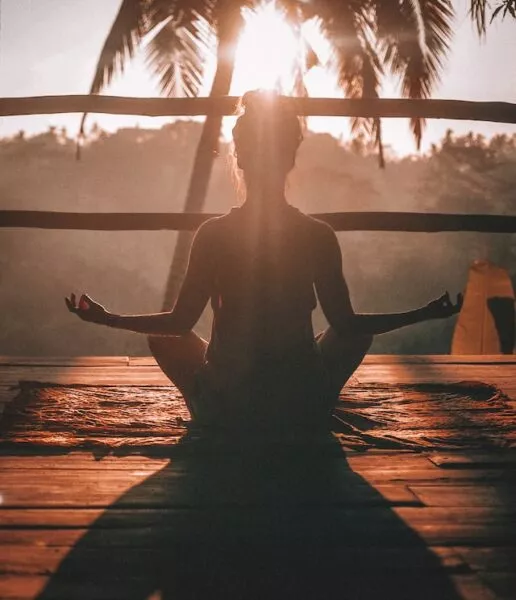 eine Person sitzt mit dem Rücken zur Kamera im Schneidersitz, die Arme seitlich angewinkelt und meditiert im Sonnenuntergang