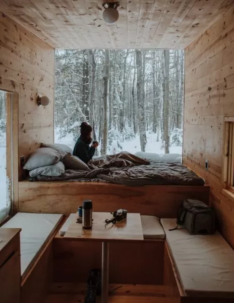 Eine Person sitzt in einem Tiny House auf dem Bett vor einer großen Fensterfront und schaut raus in eine Winterlandschaft