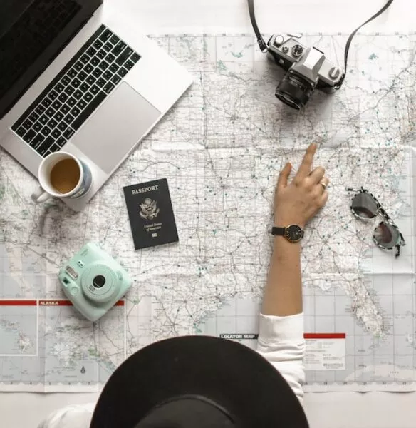 Eine Person mit Hut beugt sich über eine Landkarte, auf der auch ein Laptop, Kameras, Reisepass und Sonnenbrille liegen