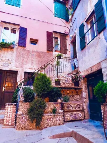 Zwei rosane Eck an Eck stehende Häuser mit vorgelagerter Treppe, die mit Pflanzen und Muscheln verziert ist 
