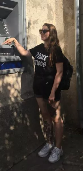 Eine Person mit Sonnenbrille, in Shirt und kurzen Hosen bedient lächelnd einen, in einer Wand eingelassenen Geldautomaten 