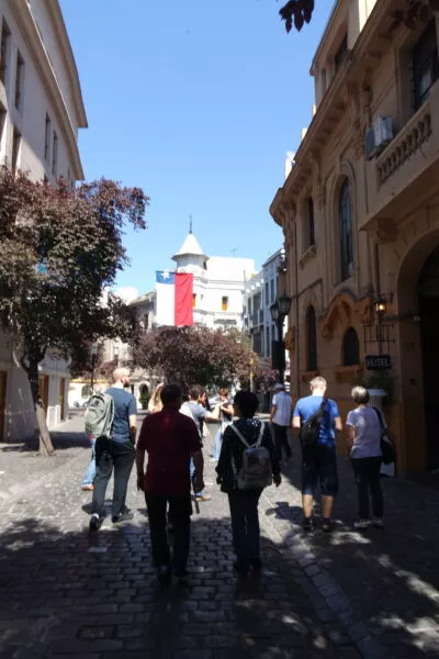 Durch eine schattige Gasse spazieren Menschen auf ein Gebäude mit chilenischer Flagge zu