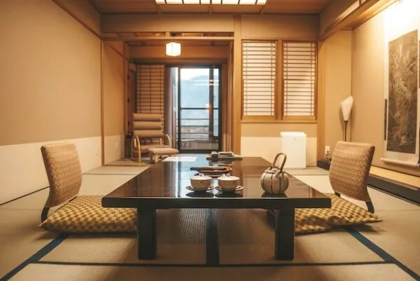 Ein japanischer Raum mit Tatami-Matten, Sitzkissen mit Lehne vor einem Tisch, Papierelementen an der Wand und einem Fenster an der äußeren Wand