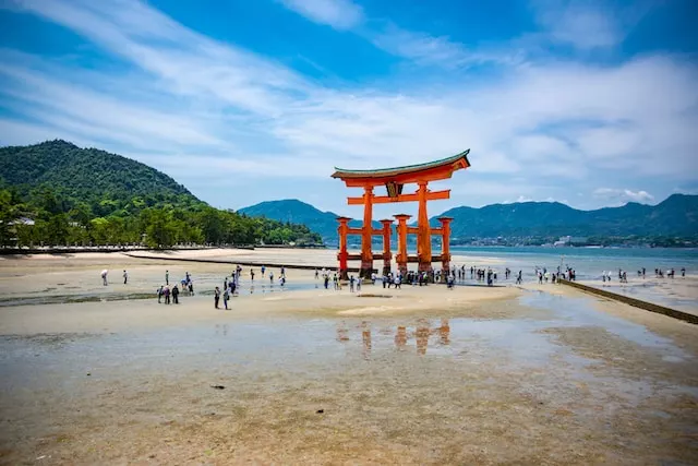 Miyajima: Ein rotes Tor auf einem Sandstrand, zu dessen Füßen viele Touristen umherlaufen. Im Hintergrund blauer Himmel und Wälder entlandg der Küste