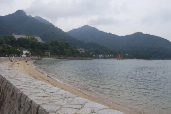 Miyajimas Küstenstreifen, in der Ferne ist ein rotes Tor mitten im Wasser zu erkennen, dahinter türmen sich Berge auf