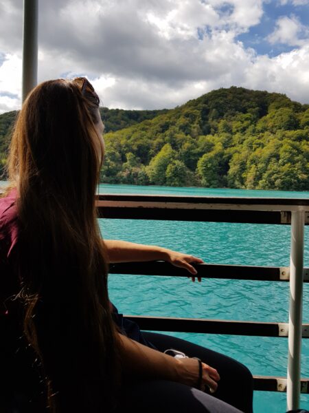 Eine Person sitzt am Rand einer Fähre und sieht auf türkisblaues Wasser der Plitvicer Seen hinaus