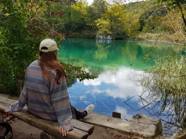 Eine Person mit Kappe sitzt auf einem Baumstamm vor einem See, der in grün und blau schimmert. Drumherum steht dichtes Gestrüpp.