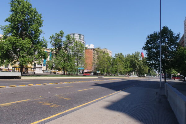 Eine breite Hauptstraße, mit vielen Bäumen und ein paar Gebäuden zur rechten und linken Seite. Im rechten Bildrand fällt ein großer Schatten auf den sonst so sonne-bestrahlen Asphalt.