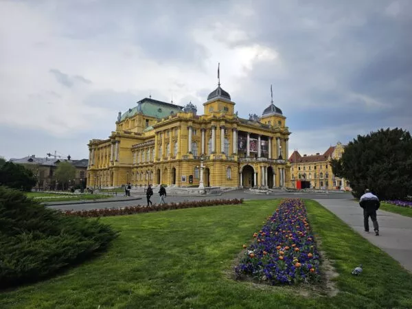 Das Nationaltheater in Zagreb mit seinen kuppelförmigen Aufsätzen. Davor eine grüne Wiese mit Blumen und Büschen.