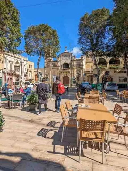 Auf einem gekachelten Platz umgeben von Gebäuden und Bäumen stehen Tische und Stühle, die wahrscheinlich zu einem Café oder Restaurant gehören