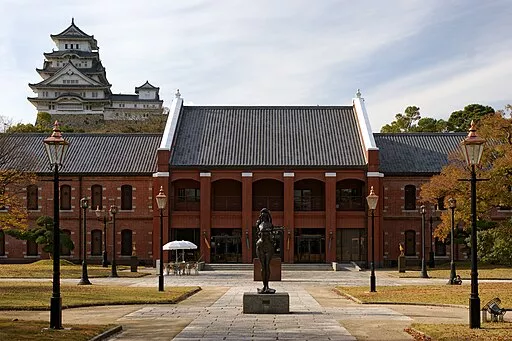 Das Himeji City Museum of Arts, ein braunes Backsteingebäude, vor dem eine Frauenstatue steht, im Hintergrund ist die Burganlage zu sehen