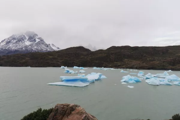 Von einer steinigen Klippe schaut man auf graues Wasser, auf dem hellblaue Eisformationen schwimmen. Im Hintergrund ein wolkenverhangener Himmel und Bergspitzen