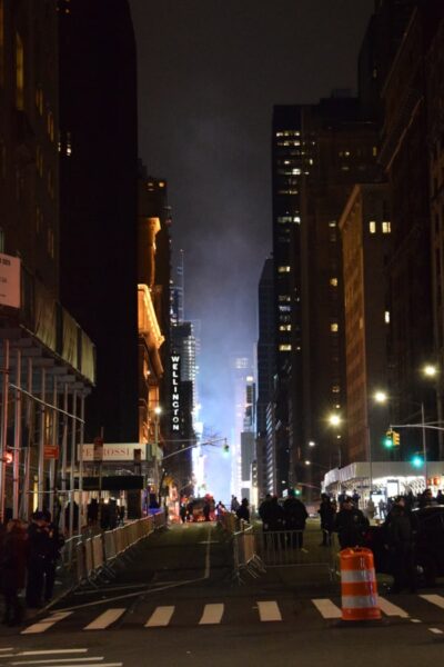 Ein abgesperrter Straßenzug in New York zu Silvester, viele Menschen sind auf der Straße, am Ende der Gasse strahlt helles Licht