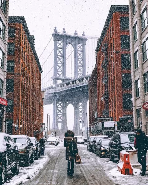 Die verschneite Brooklyn Bridge durch die Häuserfassaden hindurch fotografiert, eine Person läuft auf der Straße darauf zu, rechts und links stehen Autos