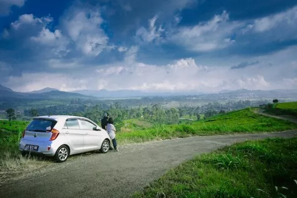 Ein kleines, silbernes Auto hält am Straßenrand, an seine Motorhaube lehnen sich zwei Personen und schauen auf die grüne Landschaft dahinter 